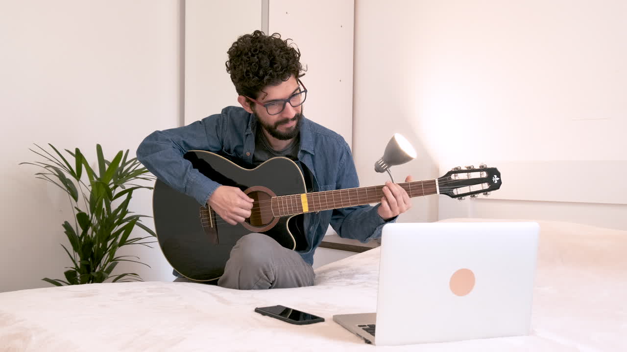 Homem sentado em casa no quarto na frente do laptop assistindo a uma aula de violão online