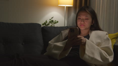 woman spending evening at home sitting on sofa with mobile phone scrolling through internet or social media 9