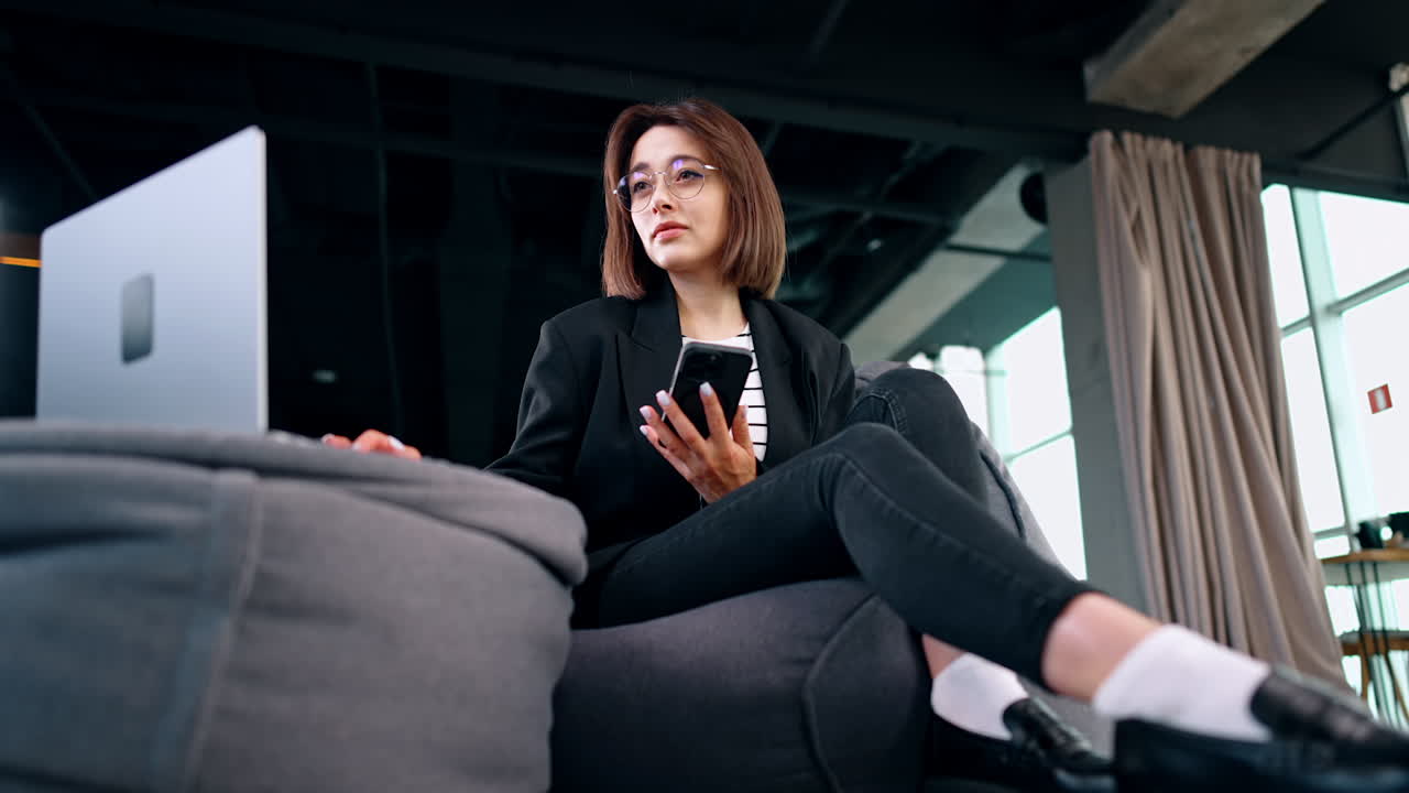 Focused lady with phone in hand looks at her laptop. Low angle view at the freelancing girl working remote.
