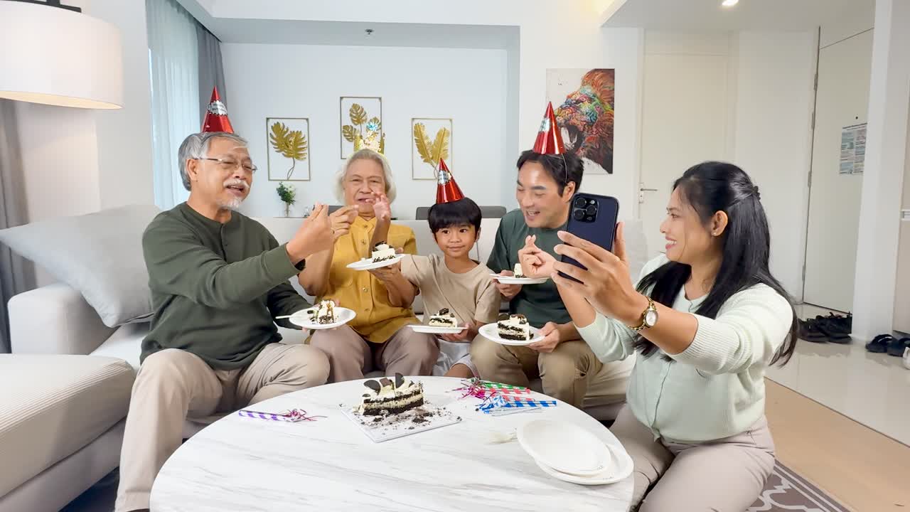 Multigenerational Asian family celebrates birthday with cake, party hats, and selfies in bright living room