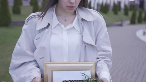 사무실 건물 앞을 걷고 있는 예쁜 여자의 클로즈업. 그녀는 물건을 상자에 담아 가지고 다닙니다. 그녀는 해고되었습니다. 그녀는 슬프다.