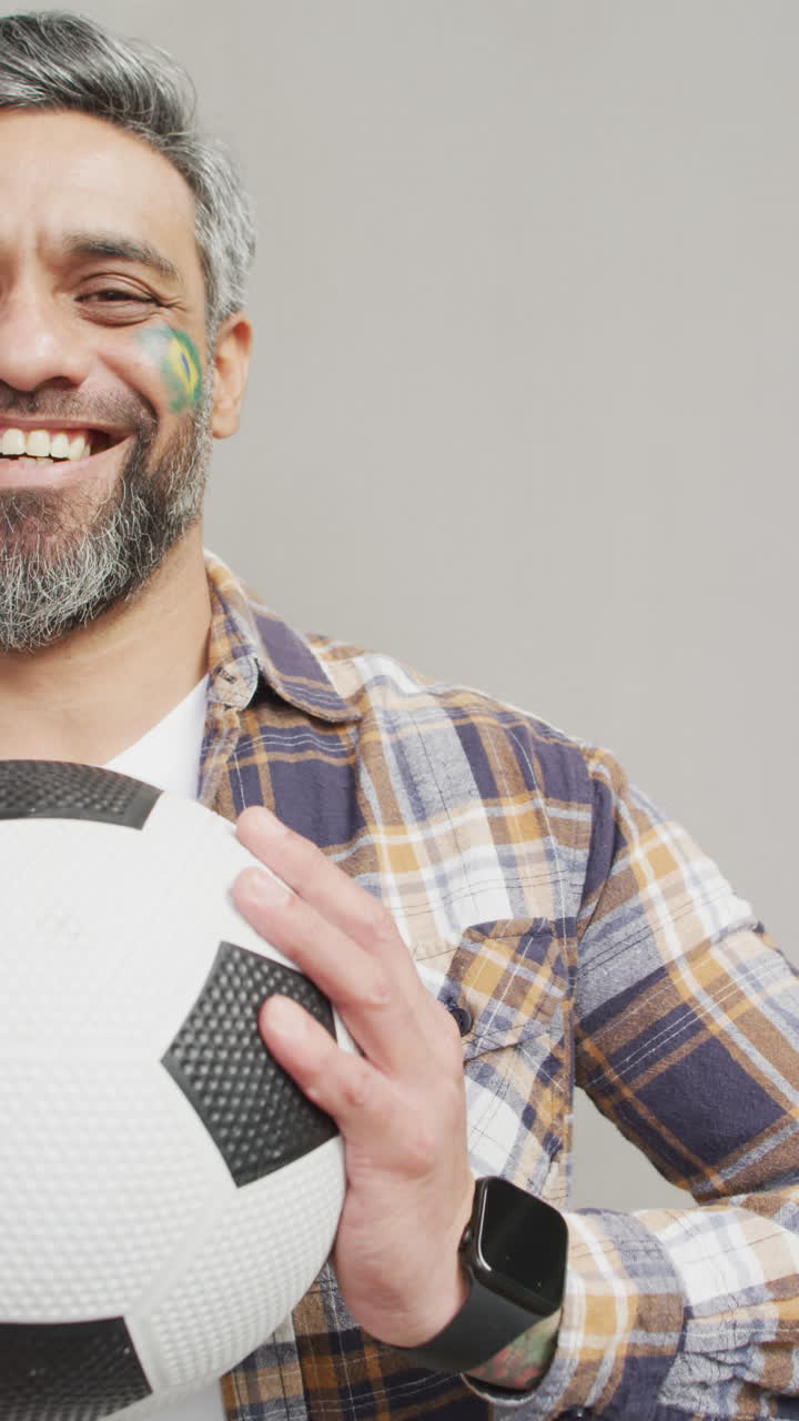 video vertical de un hombre biracial feliz sosteniendo una pelota de fútbol con la bandera de brasil en la mejilla