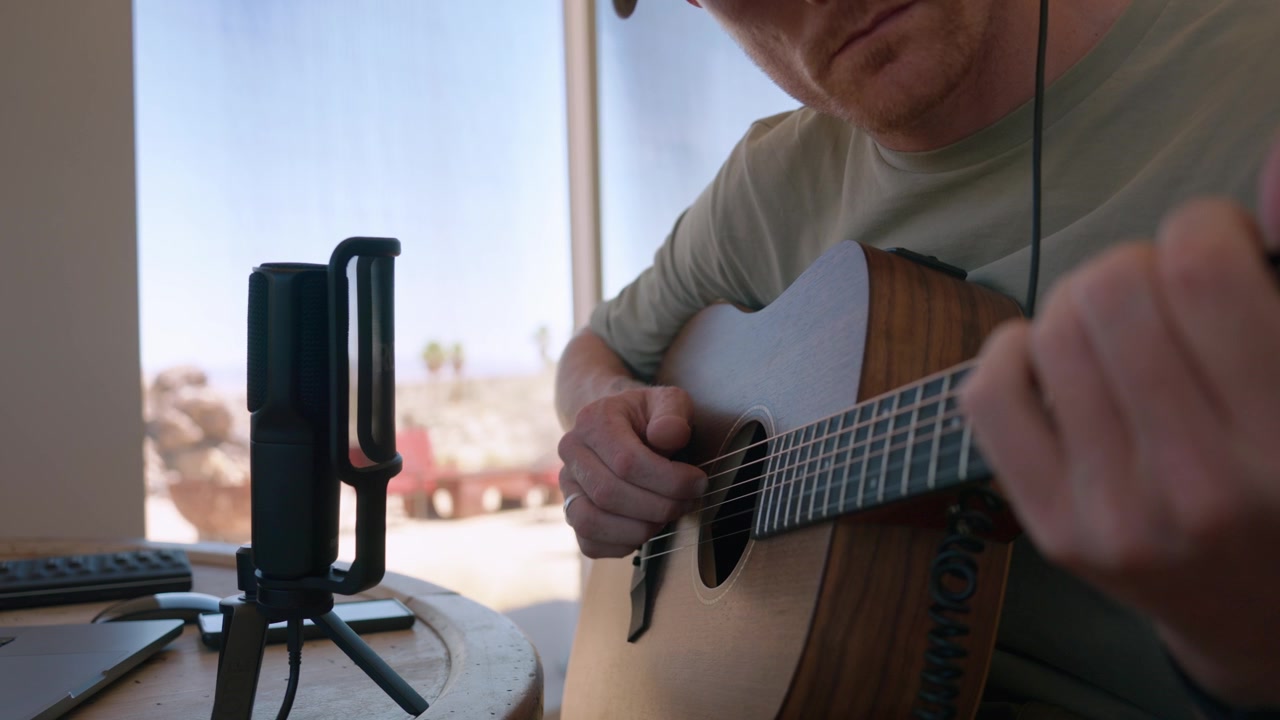 Um close-up de um homem gravando violão acústico com um microfone de estúdio ao lado de uma janela com vista para o deserto