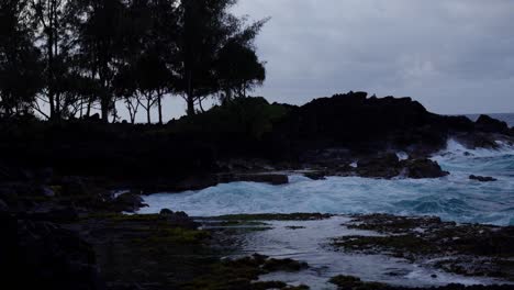 a quiet coastal scene shows swirling blue waves lapping against rugged black volcanic rocks as evening settles in, highlighting the raw and untamed beauty of the shoreline.