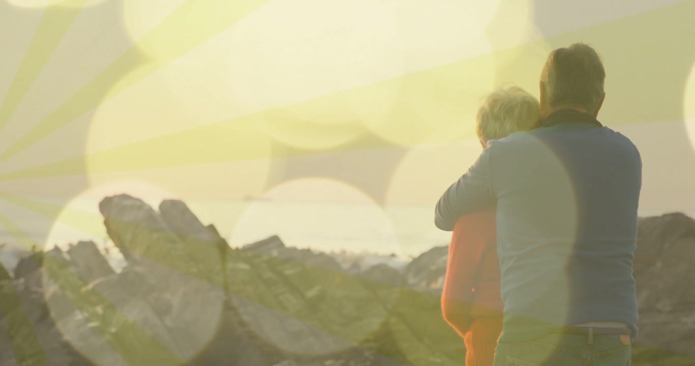 Älterer Mann mit grauem Oberteil hält Frau mit orangefarbener Jacke auf Felsen, im Sonnenlicht, unscharfer Ozean (Bokeh), mit Textfreiraum