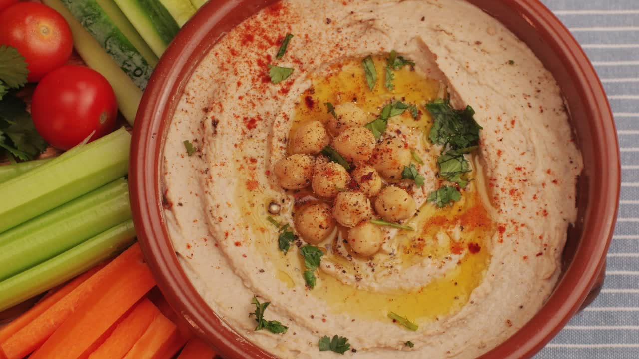 Hummus among fresh vegetables on table top view. Hummus garnished with chickpeas, olive oil, spices and greens. Hummus or fava, a hearty snack of legumes. Kosher healthy vegan food. Lean, vegetarian dish