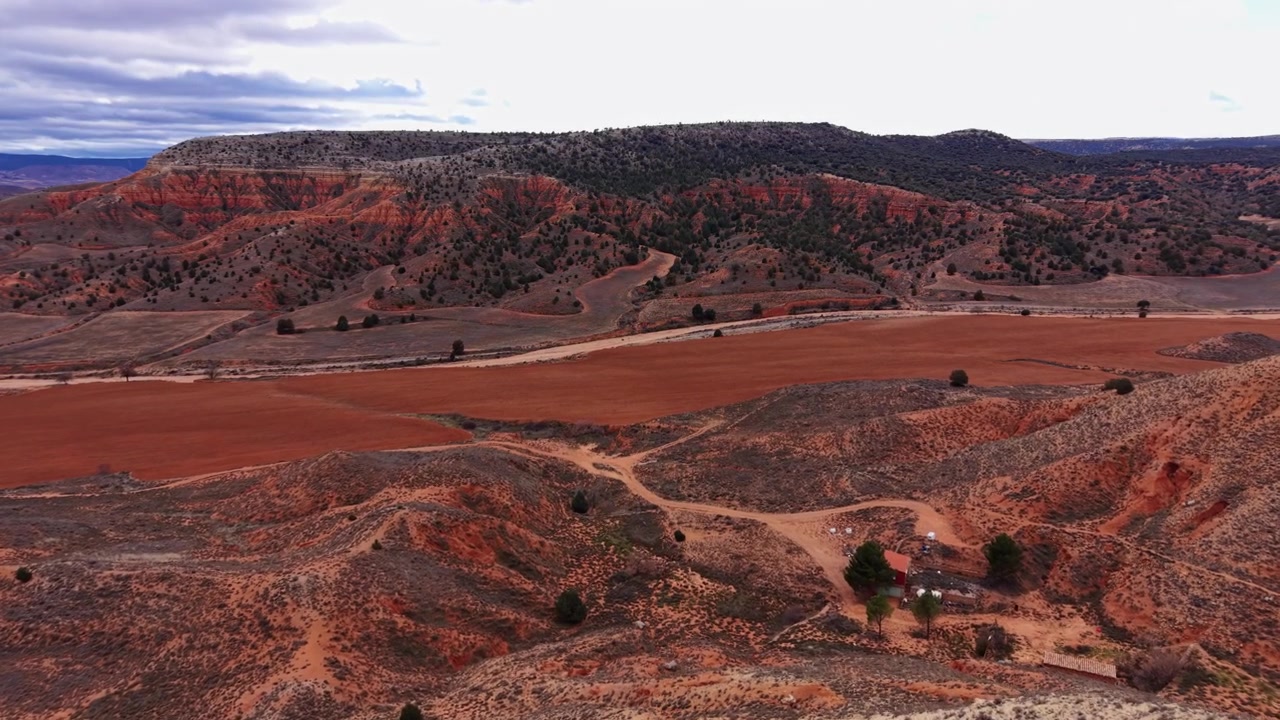 Red rock canyon landscape in the American Southwest with vast farmland, scattered trees, dirt roads, a small rural homestead, and dramatic cloudy skies over eroded mesa formations