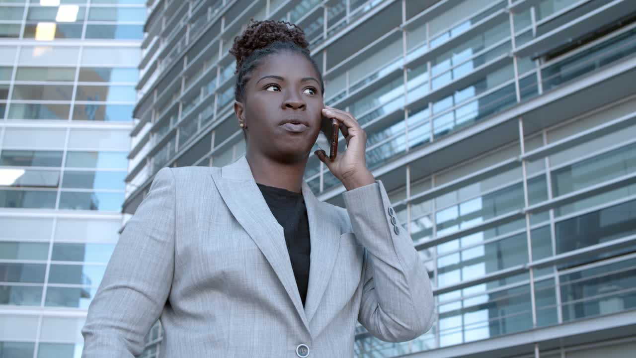 Static shot of a confident African American businesswoman standing among city office buildings and talking on mobile phone