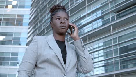 static shot of a confident african american businesswoman standing among city office buildings and talking on mobile phone