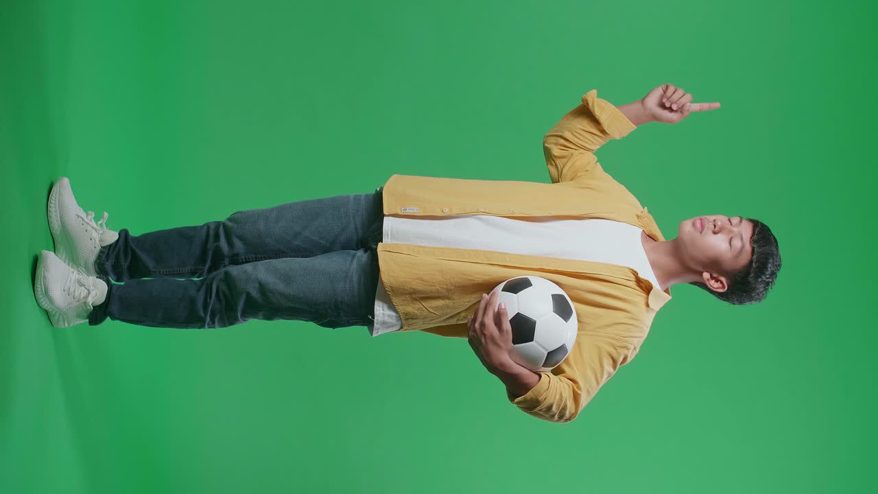 닫힌 눈의 전체 몸 측면 뷰 아시아 소년은 공을 들고 지표손가락을 올리고 머리를 흔들며 녹색 화면 배경에서 축구를 응원합니다.