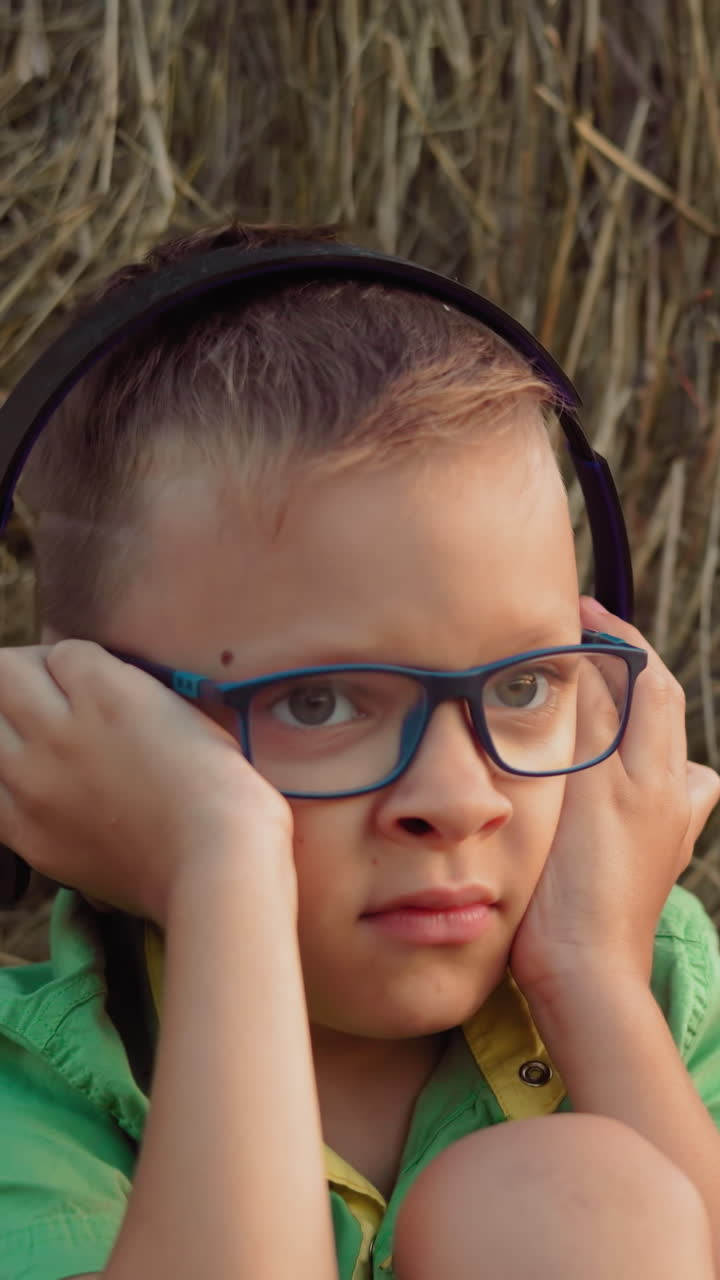 niño pequeño sentado en camisa verde ajusta auriculares mientras descansa contra una bala de heno, con manchas distintivas en sus mejillas, en un entorno rural sereno