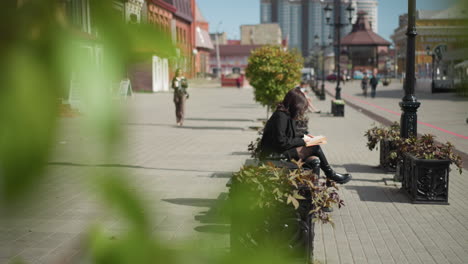 green leaves swaying in the wind with woman seated cross-legged reading book on urban walkway, blurred background features potted plants, passing cars, pedestrians and buildings