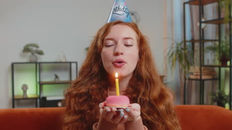 happy woman wears birthday hat makes wish congratulating blowing burning candle on cake at home