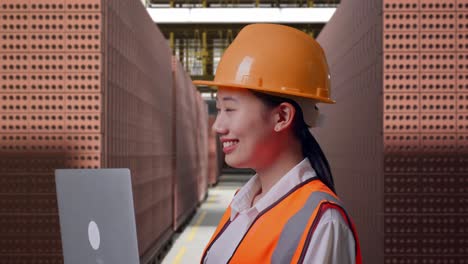 Close Up Side View Of Asian Female Engineer With Safety Helmet Working On A Laptop And Looking Around While Standing With Red Brick Packed in Stacks Are Stored