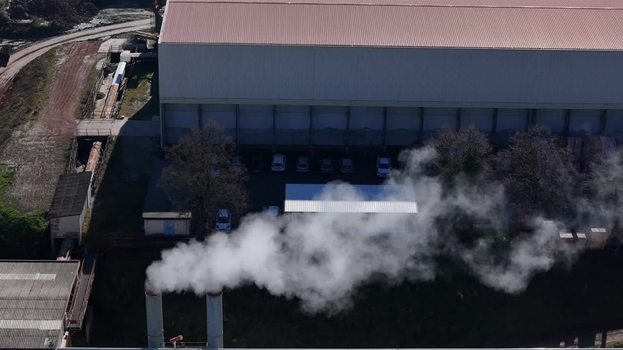 Ripresa aerea in allontanamento di un impianto industriale con ciminiera che emette fumo e vapore sopra i tetti delle fabbriche, evidenziando l'attività industriale e l'inquinamento ambientale