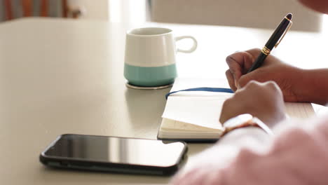 African-American-woman-is-writing-in-a-notebook-with-a-smartphone-and-a-mug-nearby,-at-home