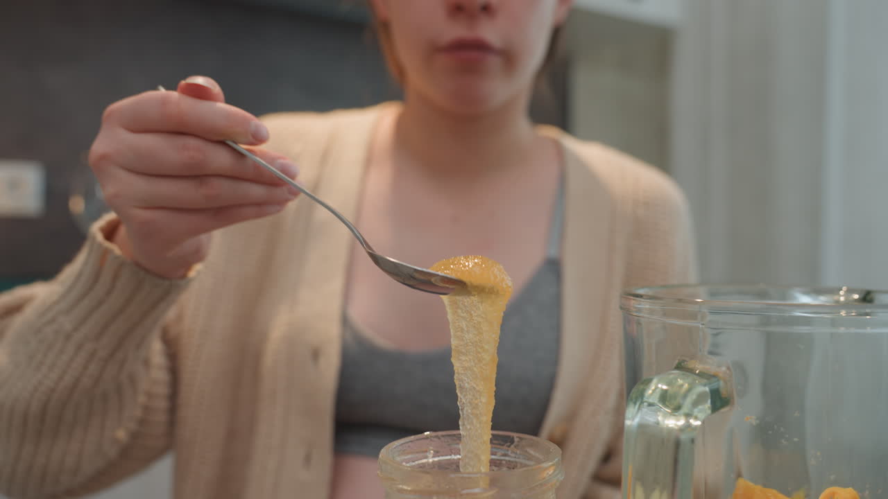 Pregnant woman scooping golden honey spoon. Closeup focus on sticky drizzle and viscous texture, jar and glass on countertop, soft cardigan, intimate sensory moment, natural remedy and prenatal taste
