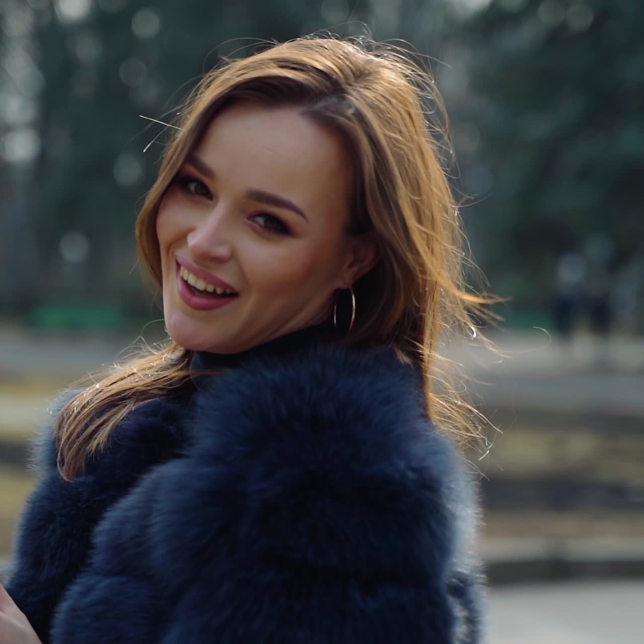 Smiling model with coffee outdoors. Beautiful girl in fur coat drinks coffee from a plastic cup while standing in the park. Young woman posing on camera. Slow motion.