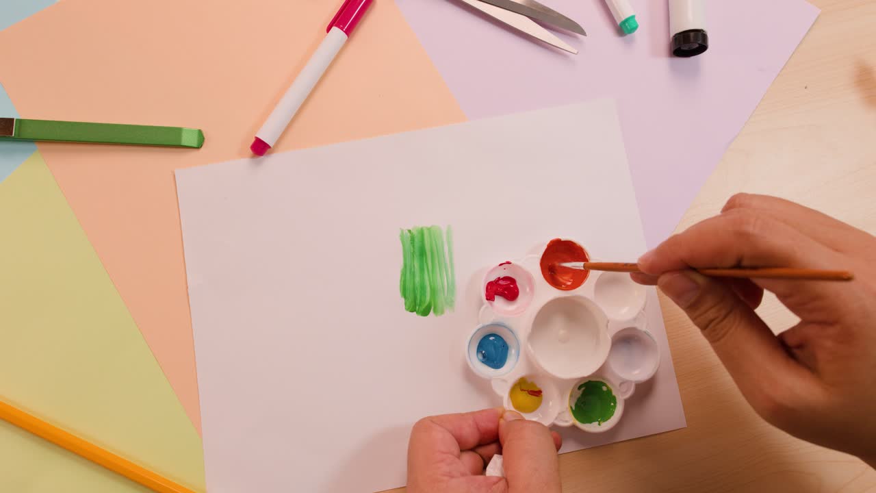 Person mixes orange paint with brush above white paper, colorful stationery, and pastel sheets
