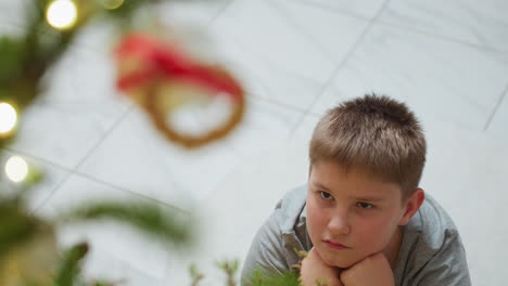 little boy sitting with two hands under chin staring at decorated christmas tree with blurred ornaments and glowing lights, peaceful indoor atmosphere, thoughtful expression during cozy festive