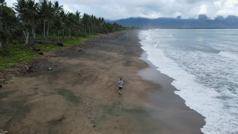 panoramic aerial follow shot of a young man running along brown sand coastline with crashing waves at tropical island in the philippines