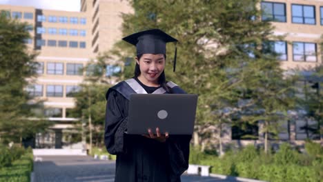 asian woman student graduates in cap and gown looking at a laptop in her hand then screaming goal celebrating in front of a magnificent university building