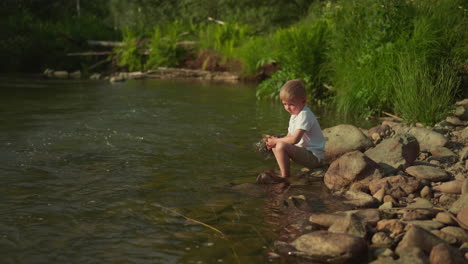 little boy plays with pebbles resting on river bank on sunny day. thoughtful child sits on big rock thinking about summer vacation. concept of recreation and childhood