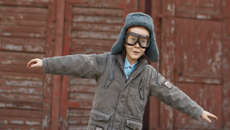 Smiling-Little-Red-Haired-Boy-In-Hat-And-Glasses-Dreaming-To-Be-An-Aviator-While-Standing-Outdoors-With-Open-Arms-And-Pretending-To-Fly