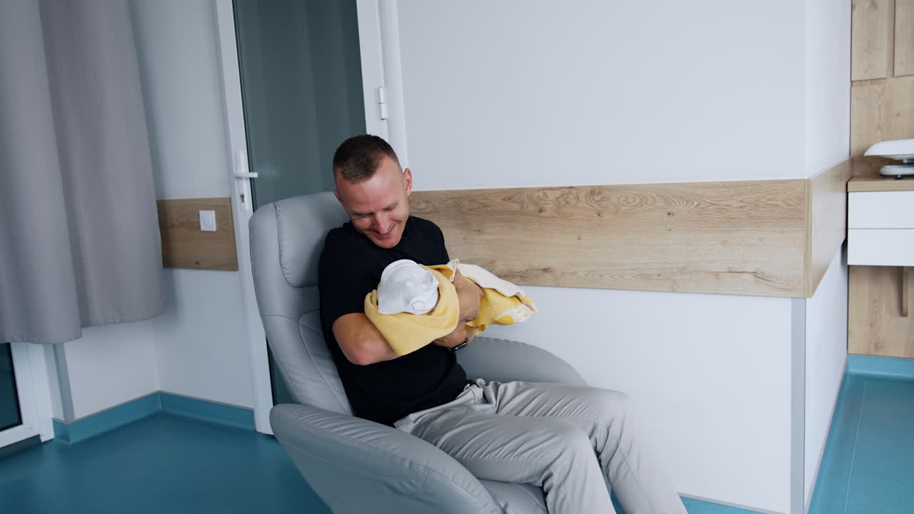 Happy smiling man sits in a chair waving a newborn. Loving and caring dad canoodling his baby.