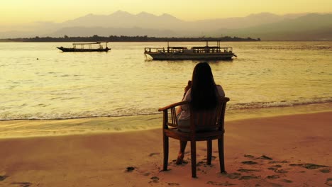 Woman-sitting-and-relaxing-in-a-chair-on-the-beach-while-taking-photos-of-the-sunset-over-the-water