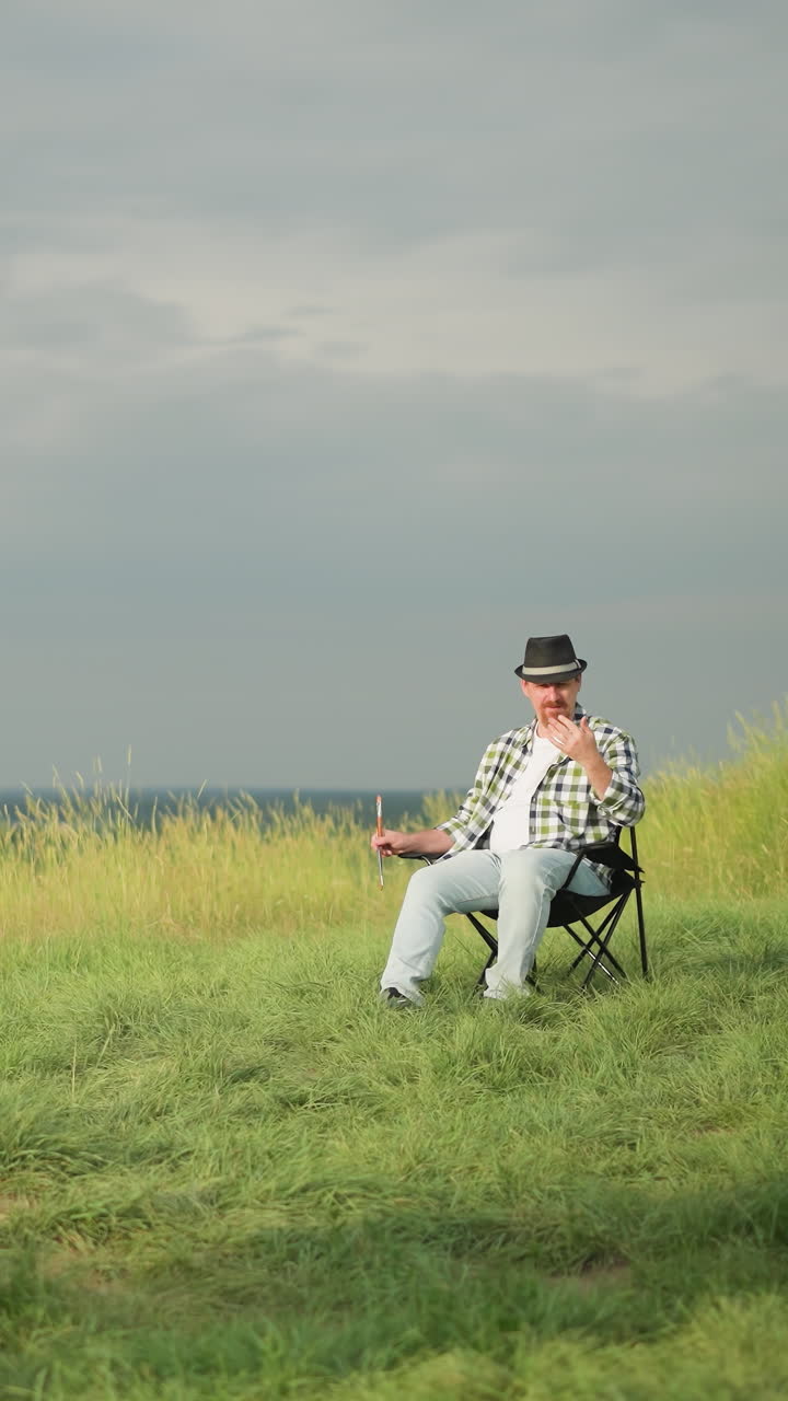 조용한 잔디에서  옷을 입은 여자가 손에 팔레트를 들고 캔버스를 칠하는 데 집중하고, 검은 모자와 체크색 셔츠를 입은 남자가 근처 의자에 앉아 있습니다.