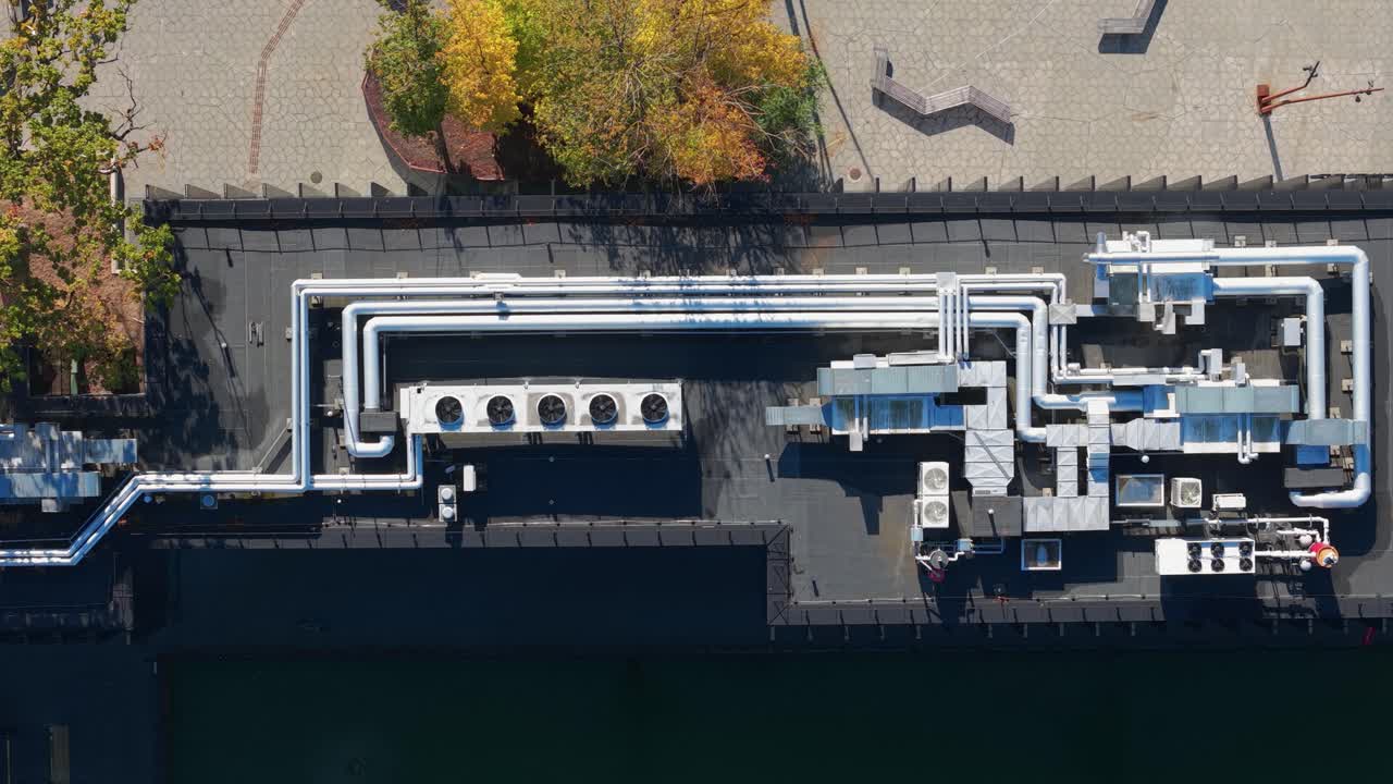 Aerial view of an industrial rooftop with visible HVAC systems, ducts, and cooling units surrounded by autumn-colored trees and an urban plaza in Kaunas, Lithuania