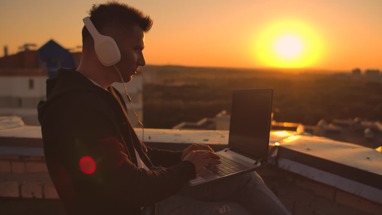 A man in a hoodie sits on the roof and listens to music with headphones typing on a laptop keyboard. Run at sunset. Freelancer works at sunset with a laptop