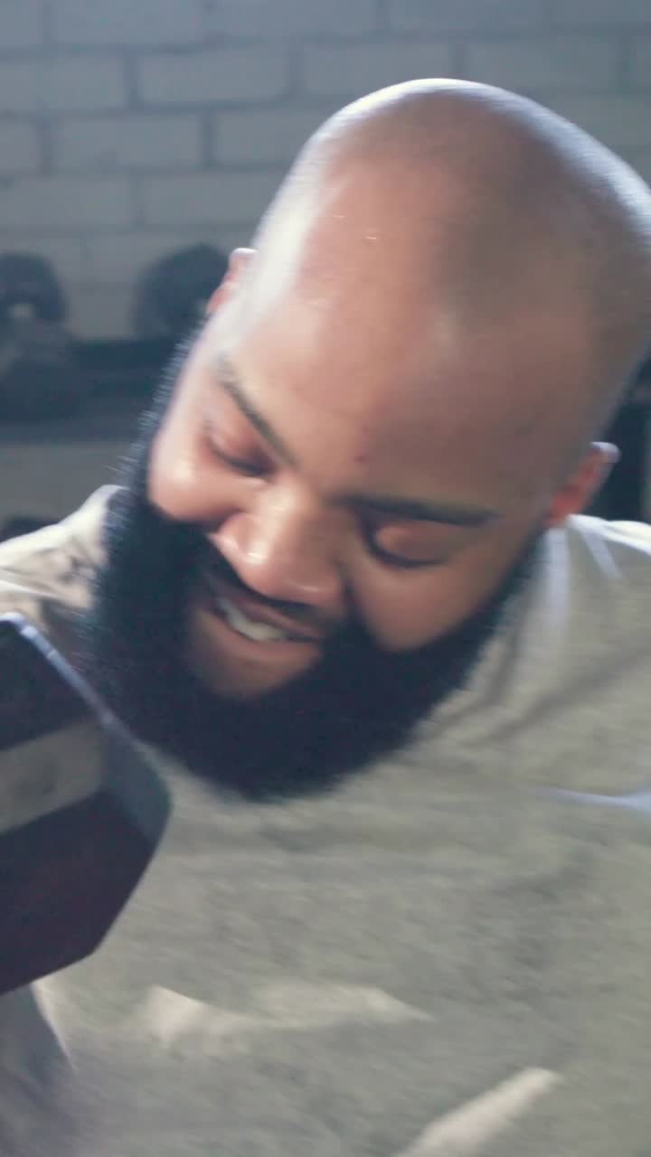 Vertical video: Camera panning, man curling hex dumbbell for camera at gym, gray tee, red shorts