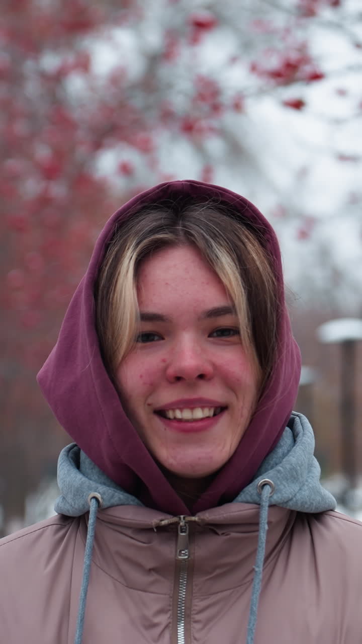 primer plano de una mujer joven con una chaqueta con capucha cálida, sonriendo con las manos en los bolsillos mientras está de pie en un camino nevado, figura borrosa visible en la distancia, árboles de bayas rojas y farolas