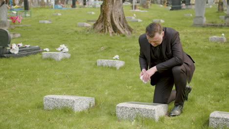 man in black suit kneeling and cleaning a tombstone with a handkerchief in a graveyard