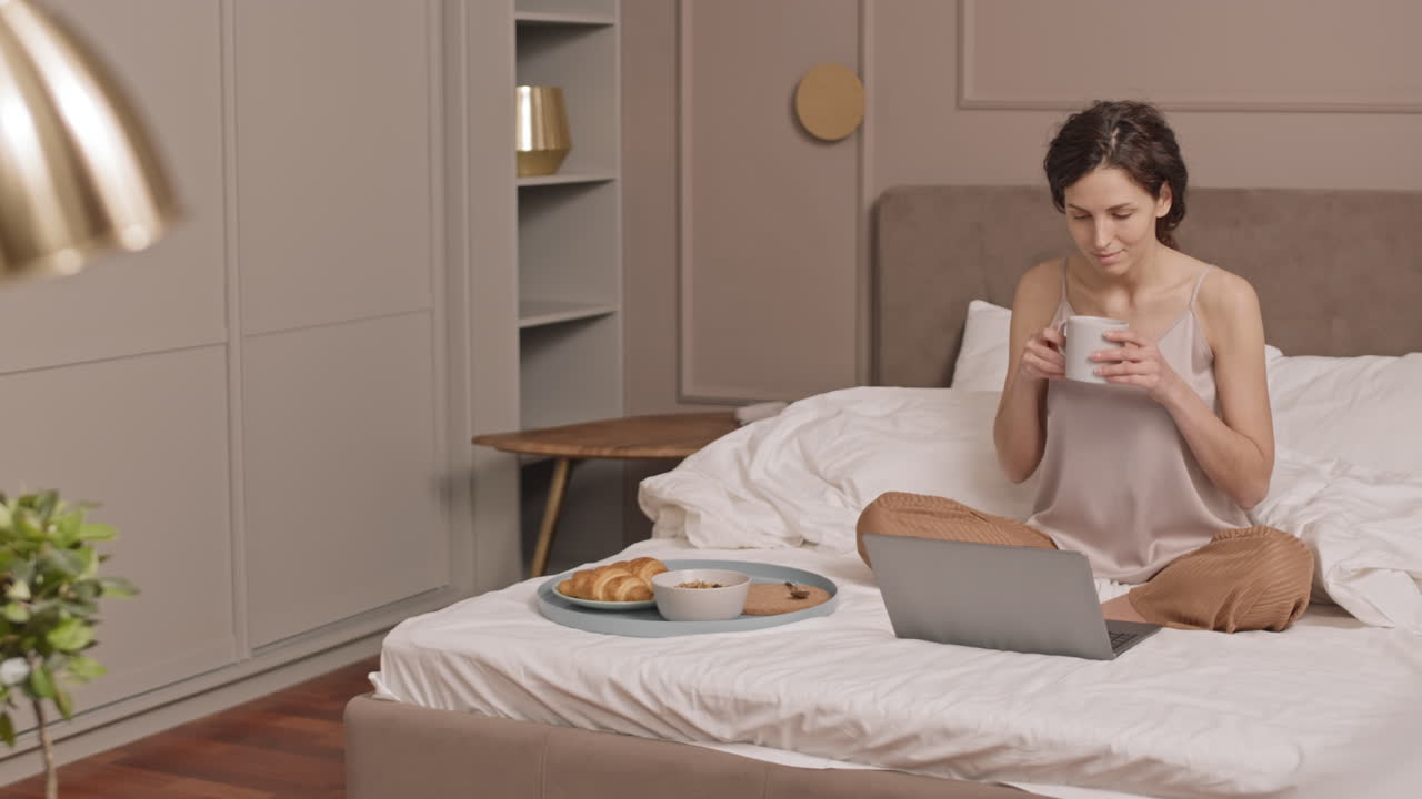 Woman enjoying breakfast in bed while working on laptop