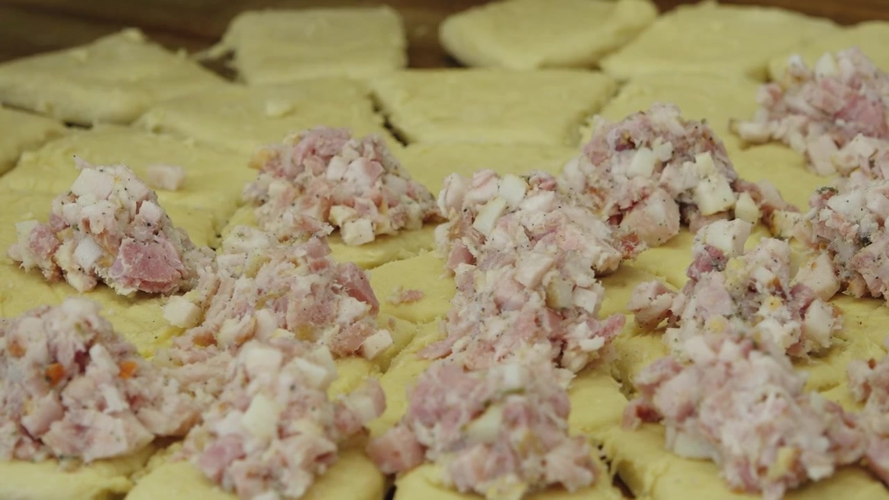 Mani guantate di nero posizionano un saporito ripieno di carne su quadrati di pasta sfoglia tagliata su una superficie di lavoro in legno durante la preparazione in panetteria.