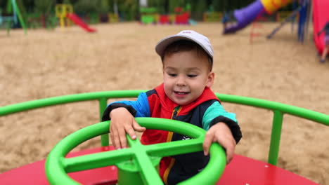 beautiful caucasian baby boy in cap sits on the merry go round. cute kid smiles to camera and jumps up happily. blurred backdrop.