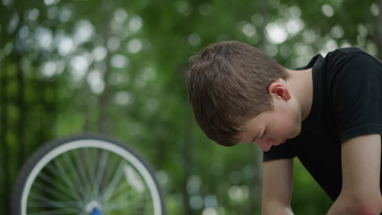 검은 옷을 입은 어린 소년은 머리를 숙여 공원 벤치에 사려 깊게 앉아 있으며, 천천히 회전하는 타이어를 가진 그의 거꾸로 된 자전거 에 조용한 배경에는 흐릿한 나무가 포함되어 있습니다.