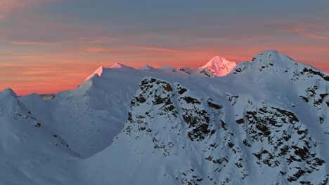 Drone-footage-capturing-the-first-rays-of-sun-illuminating-a-snow-covered-mountain,-with-the-camera-orbiting-around-a-foreground-rock