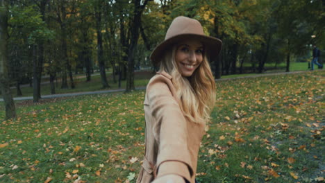 rear view of caucasian young blonde woman in a hat and coat walking in the park and leading the camera with her, then she turns and smiles