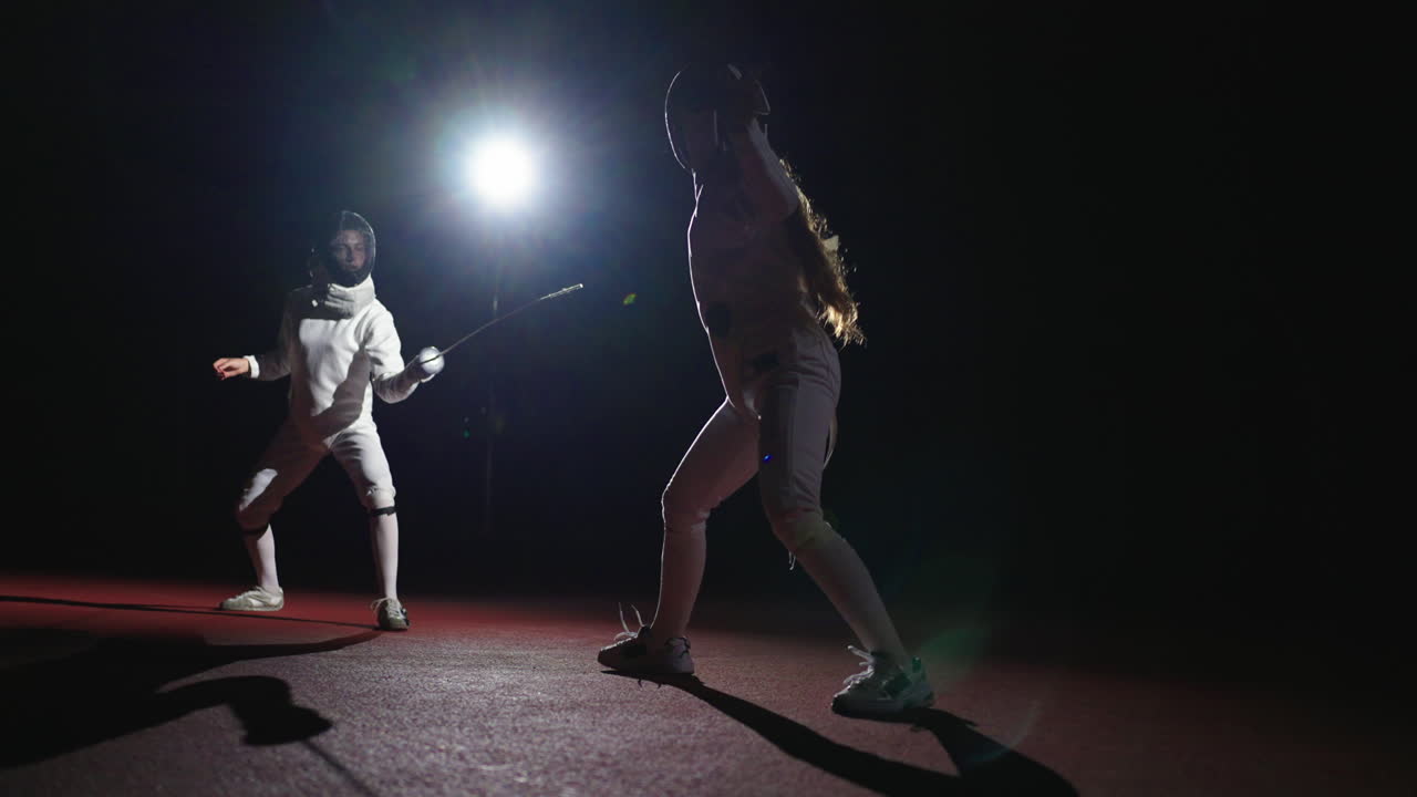 Two fencers compete in a dark arena with spotlights shining on them.