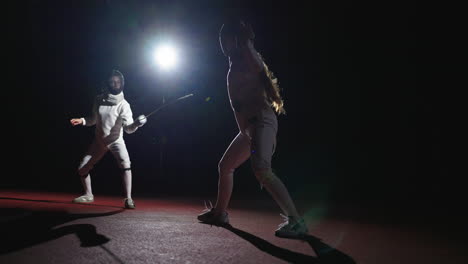 dos esgrimistas compiten en una arena oscura con los reflectores brillando sobre ellos.