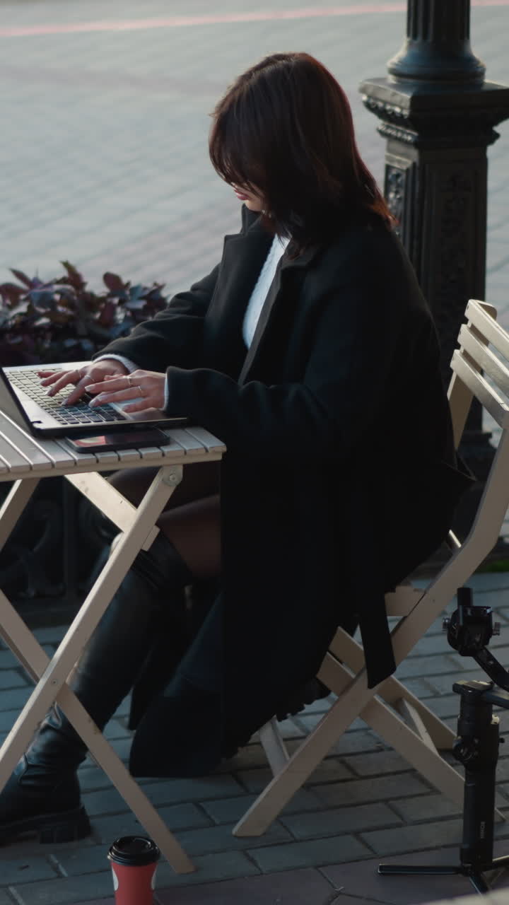 mujer profesional trabajando en una computadora portátil al aire libre en una cafetería rodeada de sillas de madera, plantas y trípode, sentada en una mesa con una taza de té, personas pasando en el fondo en una pasarela pavimentada