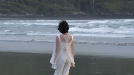 woman in white outfit walking barefoot on the beach with rough waves in the background