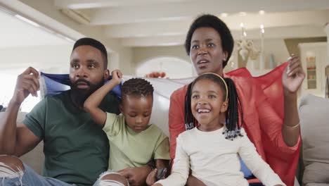 Padres-Afroamericanos-Con-Hijo-E-Hija-Viendo-Deportes-En-Televisión-Con-Bandera-En-Casa,-Cámara-Lenta