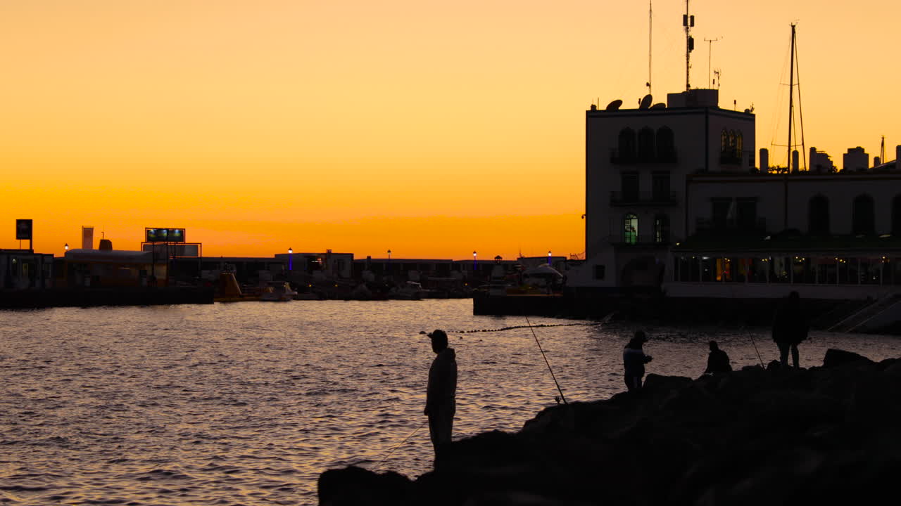 Fishermen on the stony coast during orange sunset casting fishing line on the open sea in the background is a city with a harbor and boats Grand Canary island valley 4k slow motion capture at 60fps.