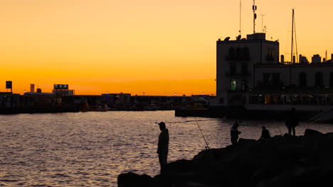 fishermen on the stony coast during orange sunset casting fishing line on the open sea in the background is a city with a harbor and boats grand canary island valley 4k slow motion capture at 60fps.