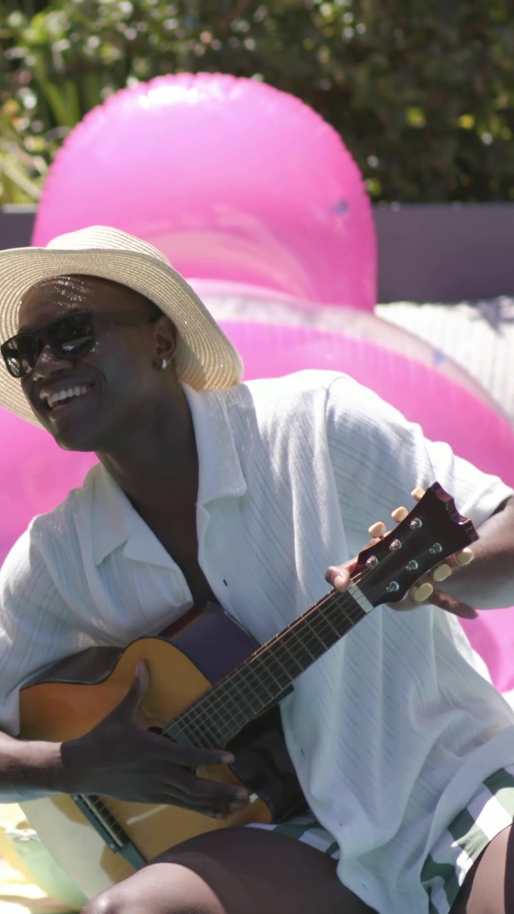 Vertikal video: Mand i stråhat spiller guitar ved poolen, inspireret af solen, med lyserøde flydere.